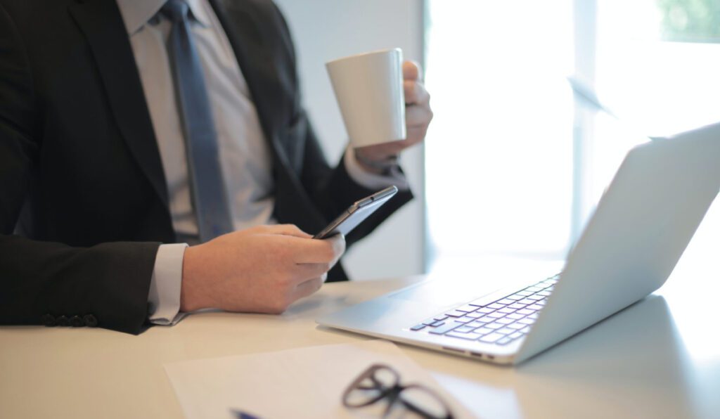 A person holding phone and mug in front of laptop